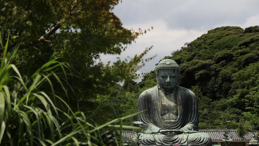 A large bronze Buddha statue among green hills, seen through tall grass