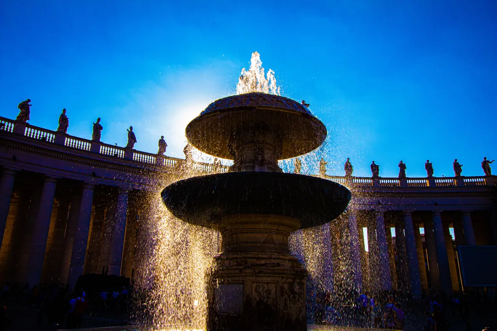 A backlit fountain in front of a colonnade topped with statues, under a deep blue sky