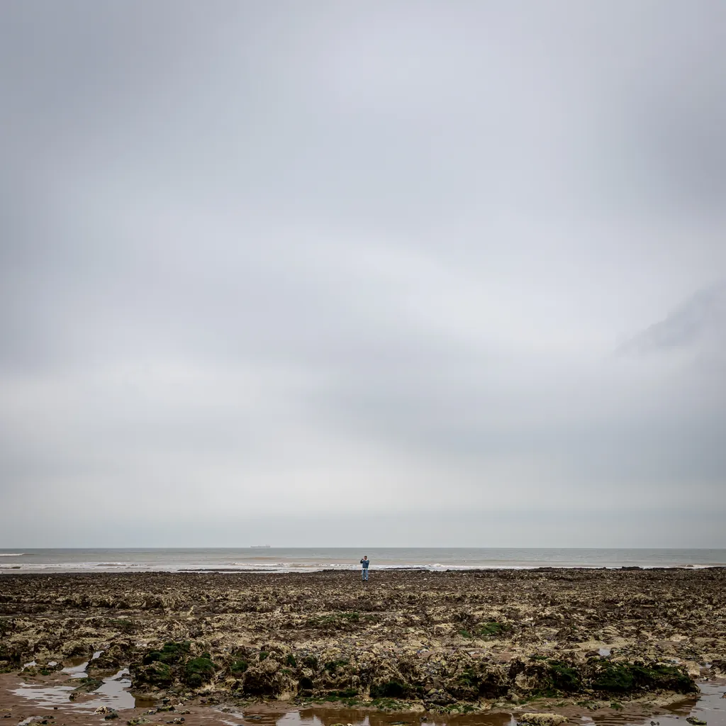 A grey sky over a rocky beach at low tide, with a small figure in the distance